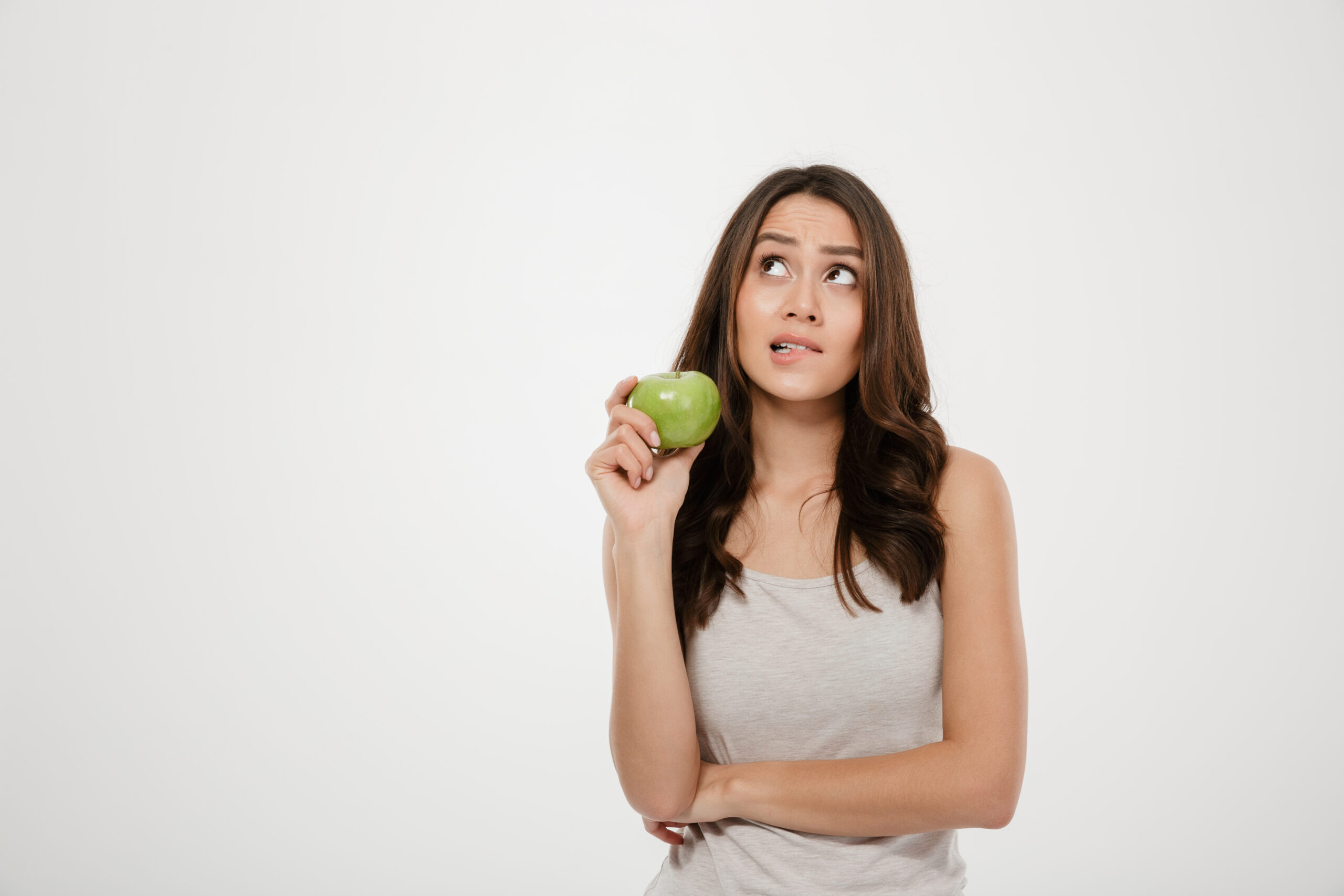 Portrait of puzzled woman looking upward holding green fresh apple thinking about healthy food isolated over white background